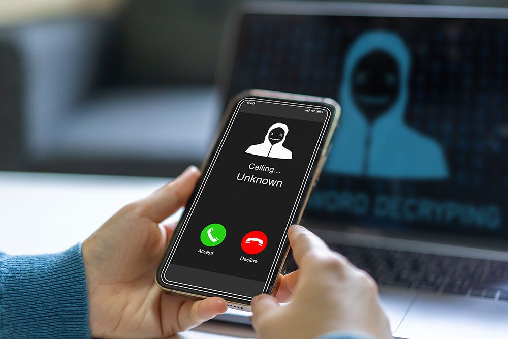 Person holding a phone showing an unknown call, with a hacker image on the laptop in the background.