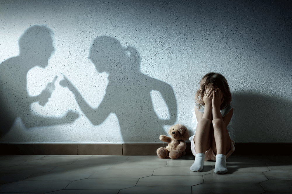 Child covering face, distressed by shadows of arguing adults on wall. Teddy bear nearby. Concept of domestic conflict impact.