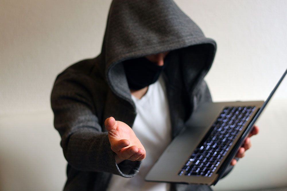 Hooded person with mask holding laptop, reaching out hand.