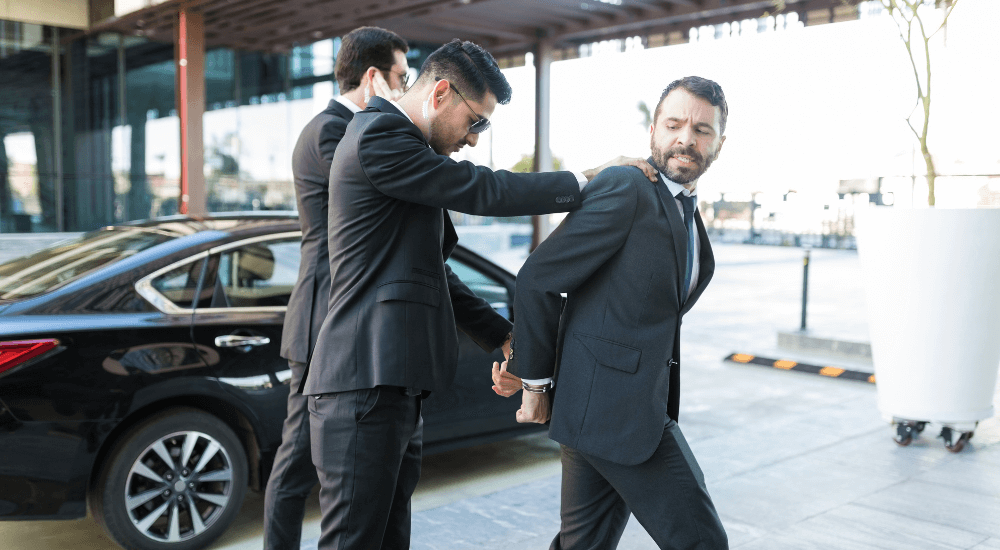 Businessman handcuffed by security near car outside office building.