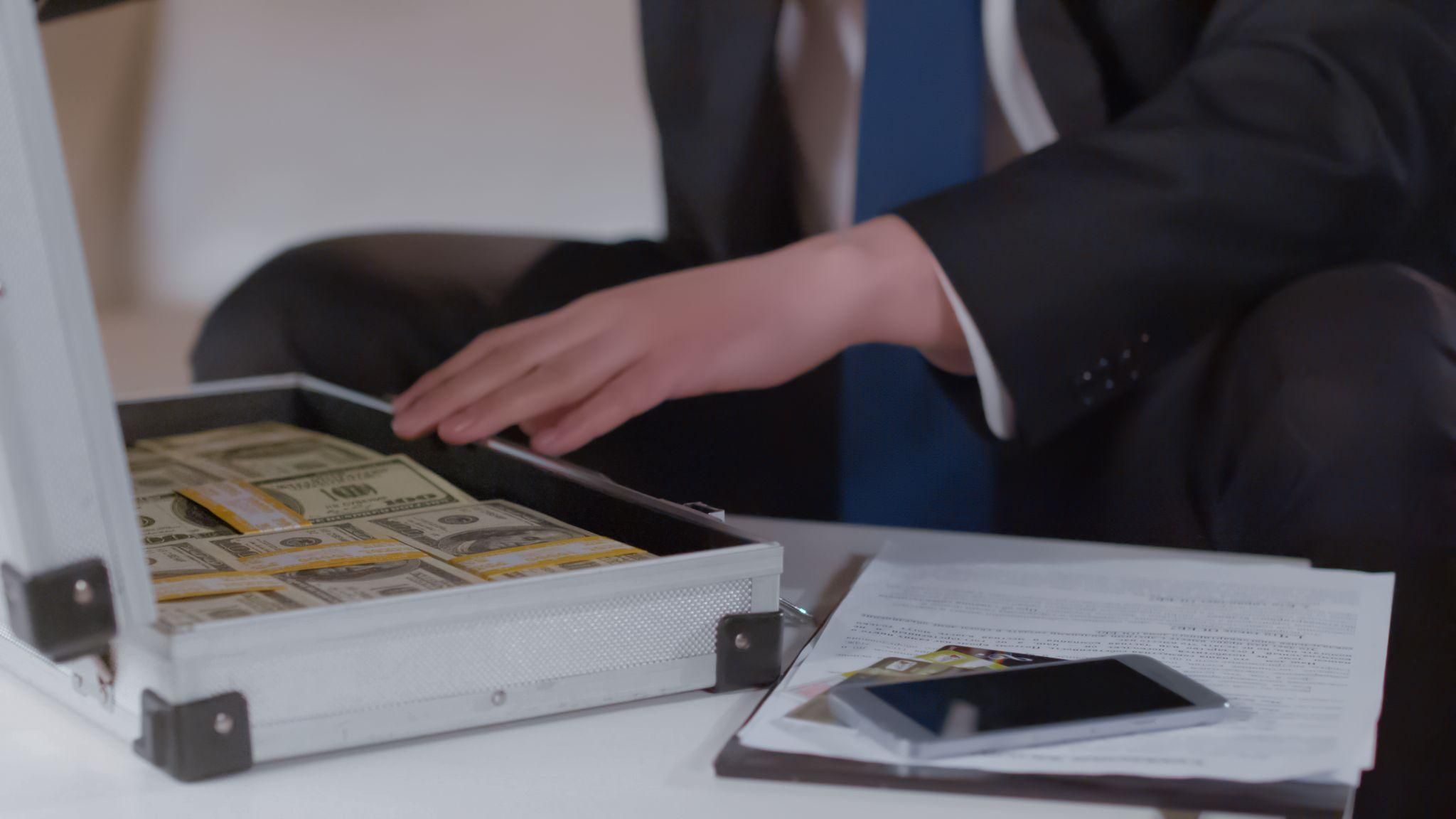Person in a suit opens briefcase full of cash beside documents and smartphone on table.