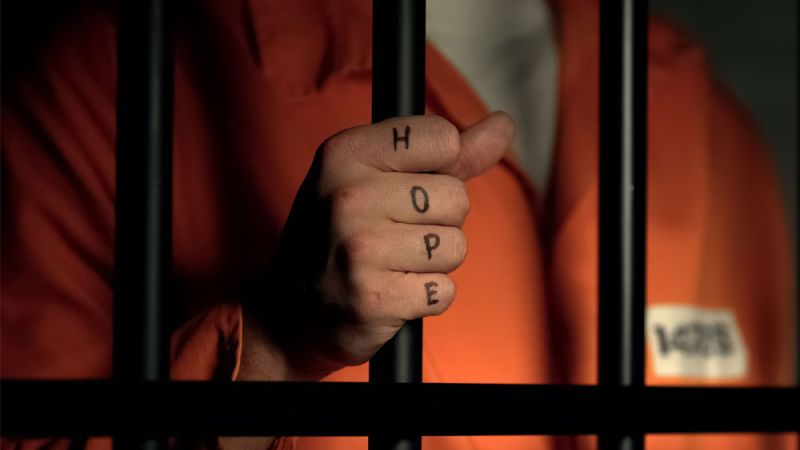 Inmate grips prison bars with Hope tattoo on fingers, wearing an orange jumpsuit.