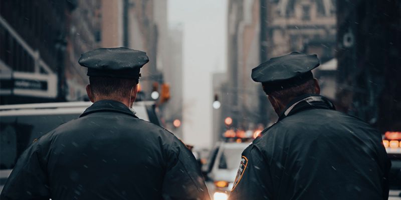Two police officers patrolling a snowy city street, facing away, with buildings and lights in the background.