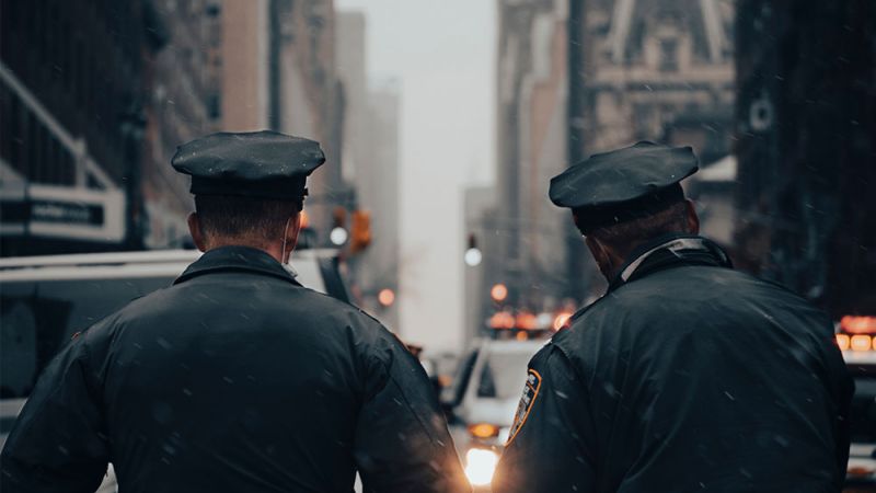 Two police officers patrolling a snowy city street, facing away, with buildings and lights in the background.