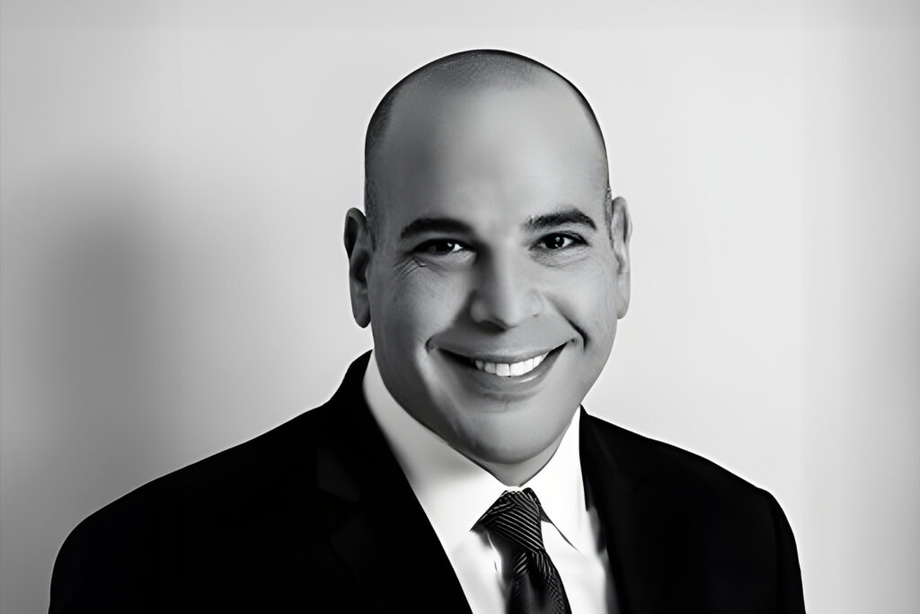 A smiling man in a suit and tie, black and white portrait against a neutral background.